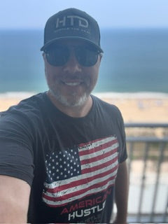A man wearing a black baseball cap with 'HTD' and 'The Hustle' logo, black sunglasses, and a black t-shirt with a distressed American flag graphic and 'AMERICAN HUSTLE' text, posing outdoors with a beach and ocean background.